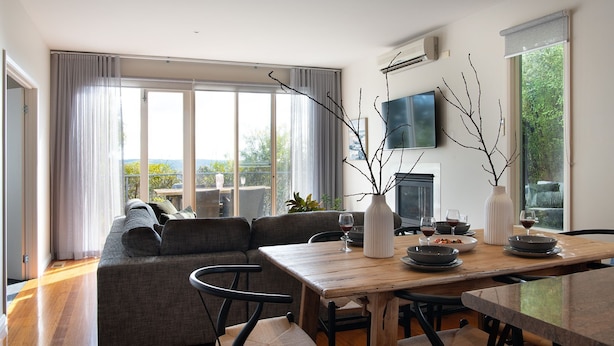 A modern living room with a sofa, dining table, and chairs. There is a fireplace, a TV mounted on the wall, and large windows with curtains. - Dahlia Daylesford - Free WIFI walking distance to Botanic Gardens with spa bath! (Daylesford)