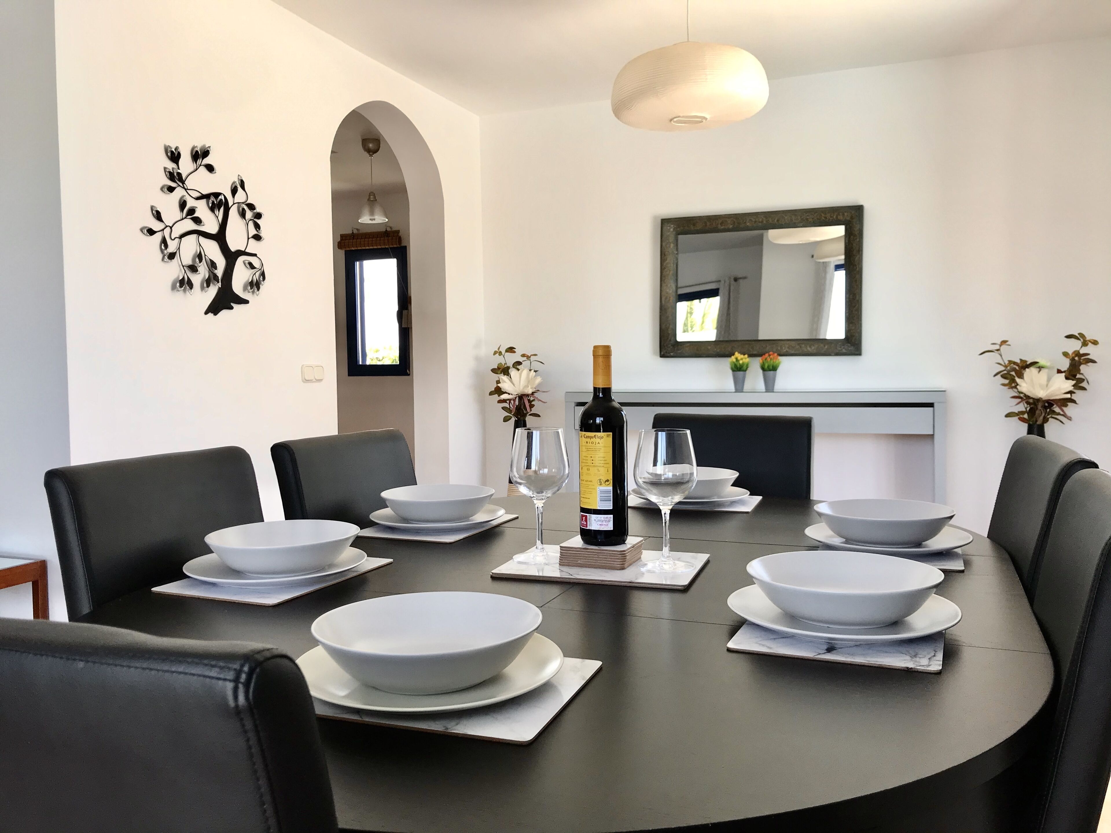 A modern dining room with a round table set for six, black chairs, white bowls, wine glasses, and a bottle of wine.