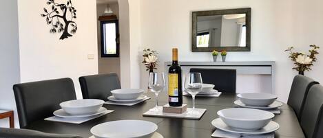 A modern dining room with a round table set for six, black chairs, white bowls, wine glasses, and a bottle of wine.