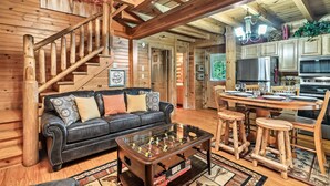 Interior acogedor de una cabaña de troncos con escalera de madera, sofá de cuero, mesa de futbolín y comedor con taburetes de madera y mesa preparada para cuatro comensales.