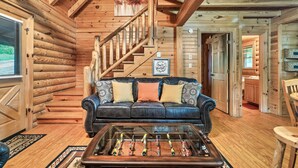 Interior de una cabaña de troncos con una escalera de madera, un sofá de cuero con cojines estampados y una mesa de fútbol.