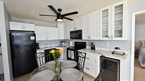 A compact kitchen with white cabinets, a black refrigerator, a microwave, and a dining area with a glass table and chairs. - Welcome To Pleasant Paradise (Fort Myers Beach)