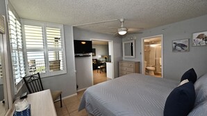 A bedroom with a bed, a desk, a chair, a ceiling fan, a window with blinds, a mirror, and a bathroom visible through an open door. - Welcome To Pleasant Paradise (Fort Myers Beach)
