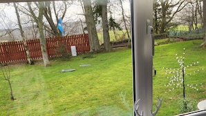 A room with a window overlooking a grassy yard, a white radiator, and a small table with a glass container. - Beautiful Studio Chalet in the Highlands (Inverness)