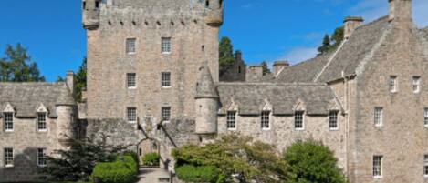 A historic stone building with multiple towers and arched windows, surrounded by greenery.