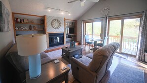 A living room with a sofa, a coffee table, a lamp, a bookshelf, a fireplace, and a large window with curtains. - Shanty Creek/ Schuss Close to lakes boat parking. golf course Sleeps 2 adults. (Mancelona)