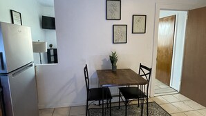 Ett kompakt kök med ett kylskåp, ett matbord med stolar och en liten matta. På väggen hänger inramade tavlor, ovanför kylskåpet sitter en tv och en trädörr leder in till ett annat rum.