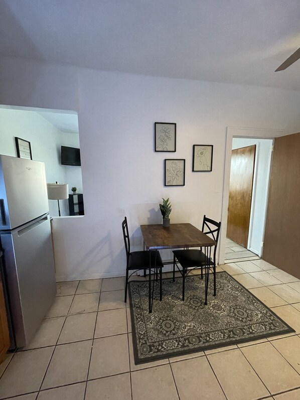A compact kitchen with a refrigerator, a dining table with chairs, and a small rug. There are framed pictures on the wall, a TV mounted above the refrigerator, and a wooden door leading to another room.