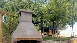 An outdoor stone-built pizza oven with a chimney, surrounded by a stone wall and a grassy area with trees. - WINTER LONG STAY.  OASIS IN CABO DE GATA (El Pozo de los Frailes)