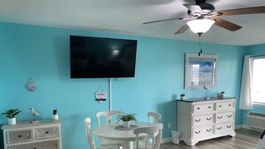 Ein Essbereich mit einem runden Tisch und weißen Stühlen, einer weißen Kommode, einem Deckenventilator und einem an der Wand befestigten Fernseher.