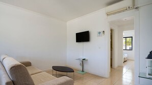 Una sala de estar con un sofá, una mesita, un televisor montado en la pared y una cocina visible a través de un vano abierto.