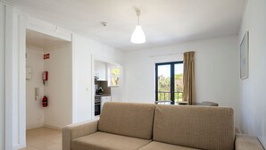 Un salón con sofá color beige, una mesa de comedor, una cocina al fondo, una ventana con cortinas y una lámpara en el techo.