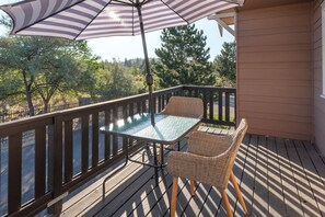 Een houten terras met een glazen tafel, rieten stoelen, een parasol en uitzicht op bomen en een weg.