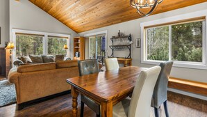 A spacious living room with a wooden ceiling, large windows, a wooden dining table, and a cozy sofa. - Private Hot Tub ~ In the heart of Sunriver ~ SHARC Passes~Fireplace~Sleeps 10 (Sunriver)