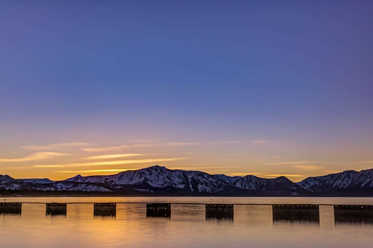 Un lago tranquilo con montañas nevadas al fondo y un puente con luces que se reflejan en el agua.