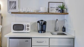 Una cocina moderna con encimera blanca, microondas, cafetera, fregadero y un refrigerador pequeño. Hay cuadros enmarcados y una planta en el estante sobre la encimera.
