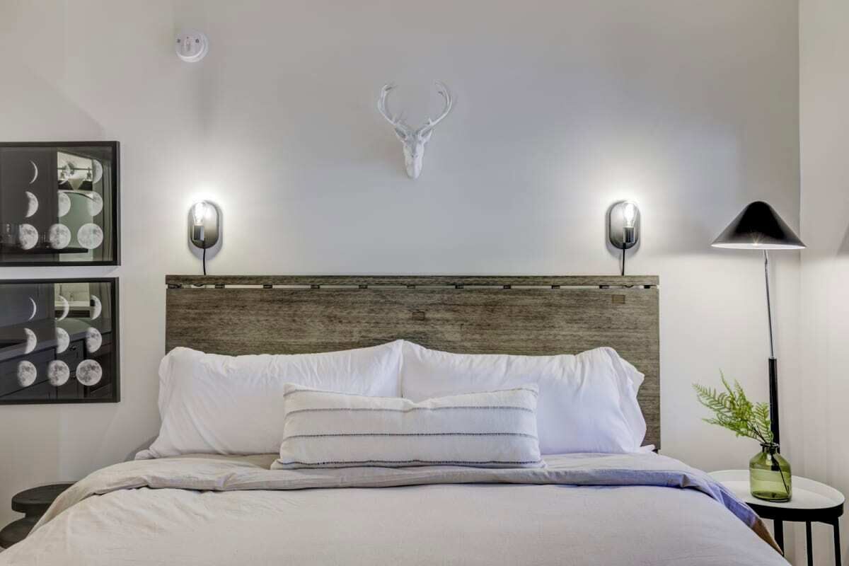 A neatly made bed with white linens and striped pillows, a wooden headboard, a wall-mounted deer head decoration, and two wall-mounted lamps.