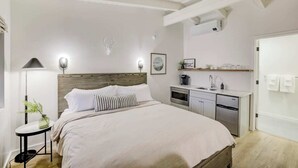 A modern bedroom with a wooden headboard, a bed with white bedding, a bedside table with a lamp, a small plant, and a built-in kitchen area with a sink and cabinets. - South Lake Chalet #10 (South Lake Tahoe)