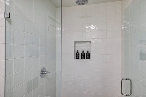 Cabina de ducha con paredes de vidrio, azulejos blancos, una ducha y tres botellas de champú y acondicionador en la pared.