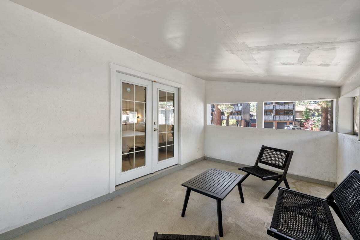 A balcony with a table and two chairs, a sliding glass door, and a view of buildings and trees.