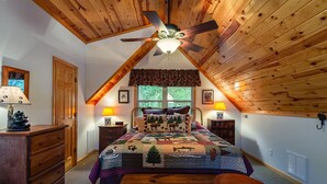 A cozy attic bedroom with a sloped ceiling, wooden beams, a bed with a forest-themed quilt, two bedside lamps, a nightstand, and a dresser. - Patriot's Point Secluded Vacation escape (Bryson City)