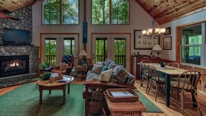 A spacious living room with a stone fireplace, a large window overlooking trees, a cozy seating area with a patterned sofa, and a dining area with wooden furniture. - Patriot's Point Secluded Vacation escape (Bryson City)