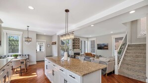 Cozinha ampla com bancada de granito, armários brancos e uma ilha central. Há uma área de refeições com um banco e cadeiras, e uma sala de estar com sofá e lareira.