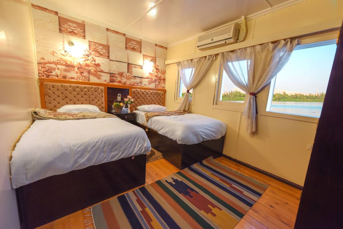 A hotel room with two beds, a large window with curtains, a patterned wall, and an air conditioning unit.