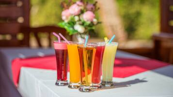 Una mesa con cuatro vasos de bebidas de colores diferentes, un mantel rojo y un florero con flores al fondo.