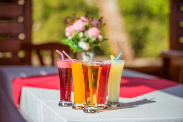 A table with four glasses of different colored drinks, a red tablecloth, and a vase with flowers in the background.
