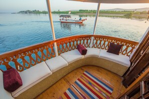 Het dek van een boot met een gebogen zitgedeelte, een uitzicht op een andere boot op het water, en een verre kustlijn met begroeiing.