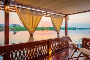 Una terraza de madera con vista a un cuerpo de agua tranquilo y vegetación al atardecer.