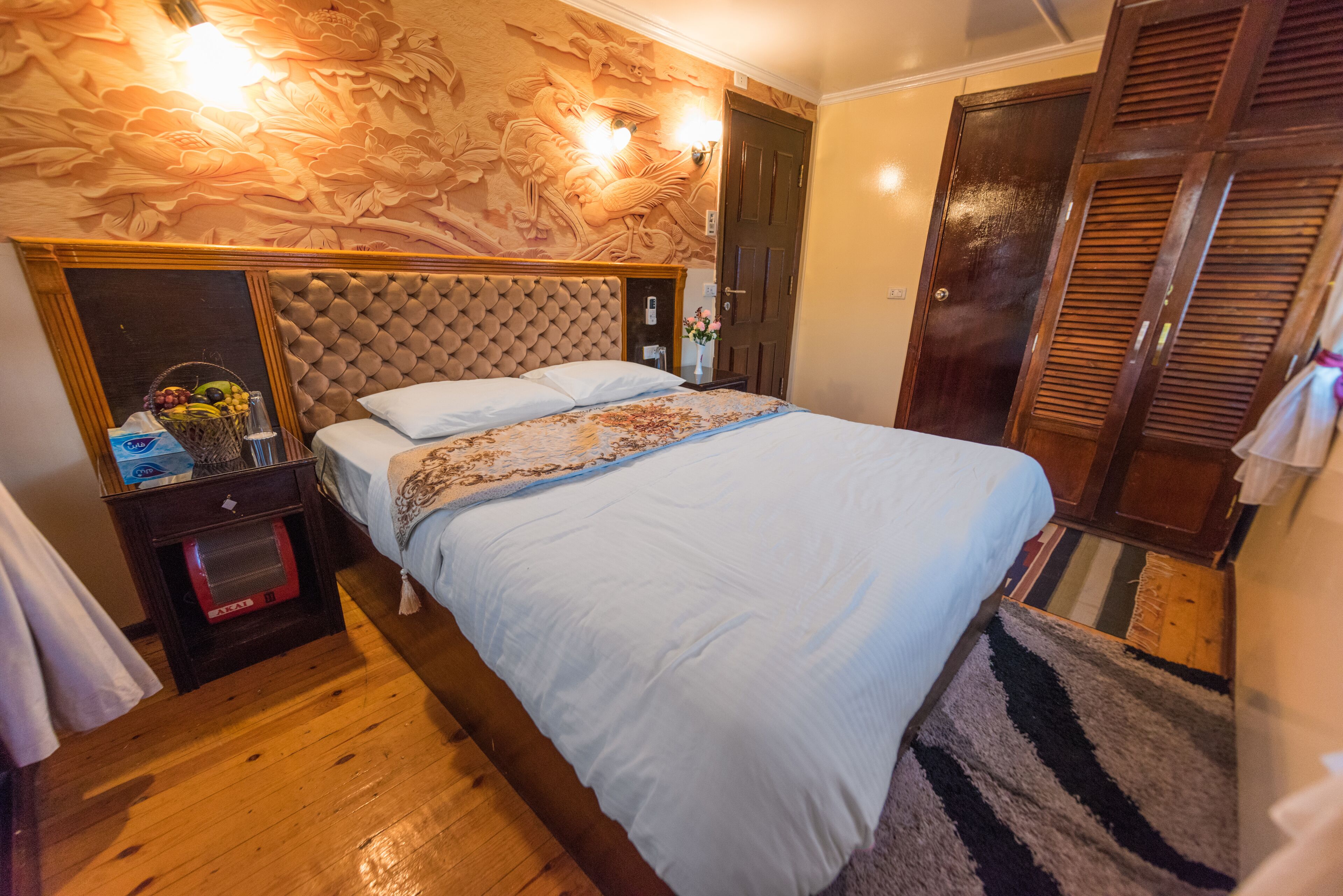 A bedroom with a large bed, a wooden headboard, a nightstand with a lamp, and a door with louvered shutters.