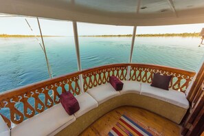 A curved deck with white cushioned seating and a colorful rug, overlooking calm water.