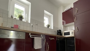 Et moderne køkken med mørke skabe, hvide bordplader og en opvaskemaskine i rustfrit stål. To vinduer giver naturligt lys, og en mikrobølgeovn er synlig på bordpladen.