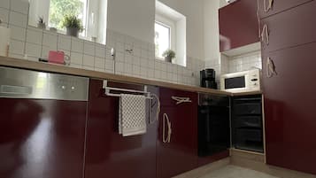 Una cocina moderna con armarios oscuros, encimeras blancas y un lavavajillas de acero inoxidable. Hay dos ventanas que proporcionan luz natural, y un microondas es visible en la encimera.