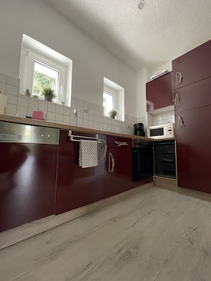 Eine moderne Küche mit dunklen Schränken, weißen Arbeitsplatten und einem Edelstahl-Geschirrspüler. Zwei Fenster sorgen für natürliches Licht, und eine Mikrowelle ist auf der Arbeitsfläche sichtbar.