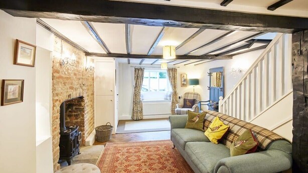 A cozy living room with exposed beams, a fireplace, a sofa with cushions, and a staircase leading to another room. - Host & Stay | Willow Cottage (Helmsley)