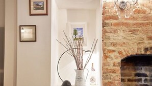 A room with a brick fireplace, a white shelf with a vase of branches, and framed pictures on the wall. - Host & Stay | Willow Cottage (Helmsley)