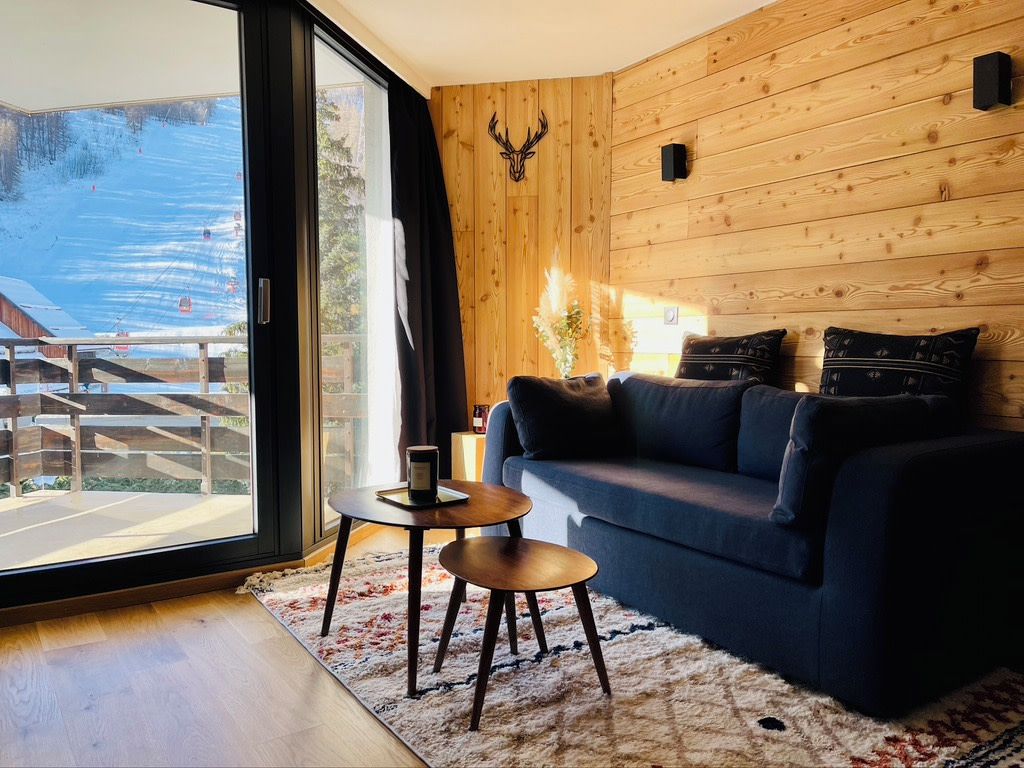 Een moderne woonkamer met een grote schuifpui naar een balkon, een donkerblauwe bank, een klein rond tafeltje en een houten wand met een opgeplakt hertenhoofd.