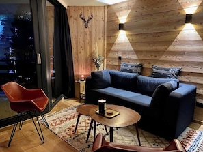 A modern living room with a sofa, a coffee table, and a red chair. There is a wooden wall with a deer head decoration and two wall-mounted lights.