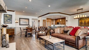A spacious living room with a brown leather sofa, a dining area with wooden chairs, and a kitchen in the background. - Highlands Slopeside #207 3 Bedroom Condo by RedAwning (Avon)