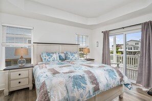 A bedroom with a bed, two bedside tables, a nightstand, a window with blinds, and a view of houses through the window. - Statuesque waterfront home by Seaside Vacations & Sales (Greenbackville)