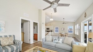 A spacious living room with a grey sofa, a striped armchair, and a glass coffee table. There is a ceiling fan, a dining area with a table and chairs, and a kitchen in the background.