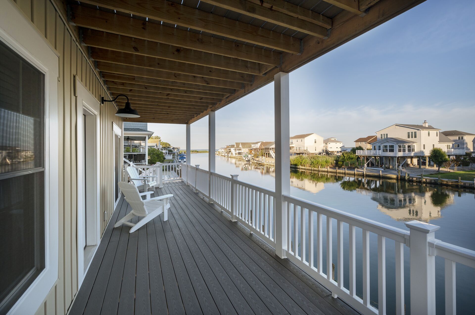 A covered wooden deck with white railings overlooking a calm water body and residential buildings.