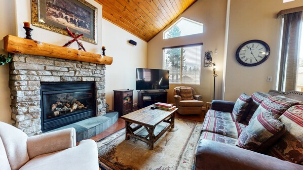 A cozy living room with a stone fireplace, a television, a sofa, and a patterned rug. - Mammoth Gateway Village 7 Pet-Friendly, Mountain Modern with Private Jacuzzi and 2 Car Garage by RedAwning (Mammoth Lakes)