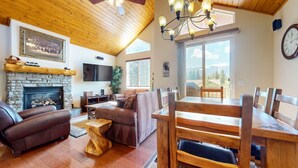 A cozy living room with a stone fireplace, leather sofas, wooden dining table, and a large window offering a mountain view. - Mammoth Gateway Village 7 Pet-Friendly, Mountain Modern with Private Jacuzzi and 2 Car Garage by RedAwning (Mammoth Lakes)