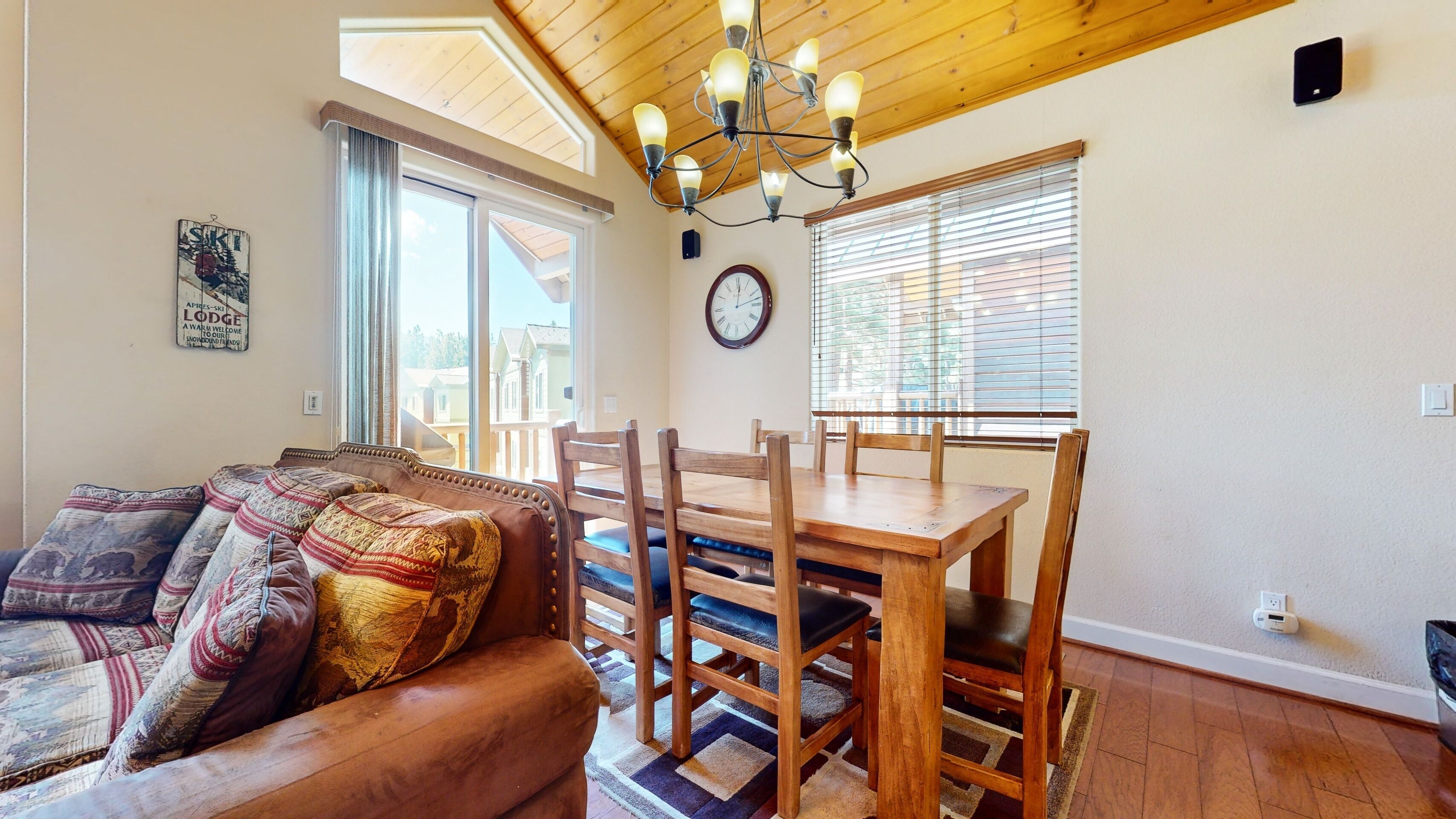 A cozy living room with a wooden ceiling, a dining table with chairs, a sofa with patterned cushions, and a large window with blinds.