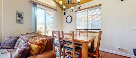 A cozy living room with a wooden ceiling, a dining table with chairs, a sofa with patterned cushions, and a large window with blinds.