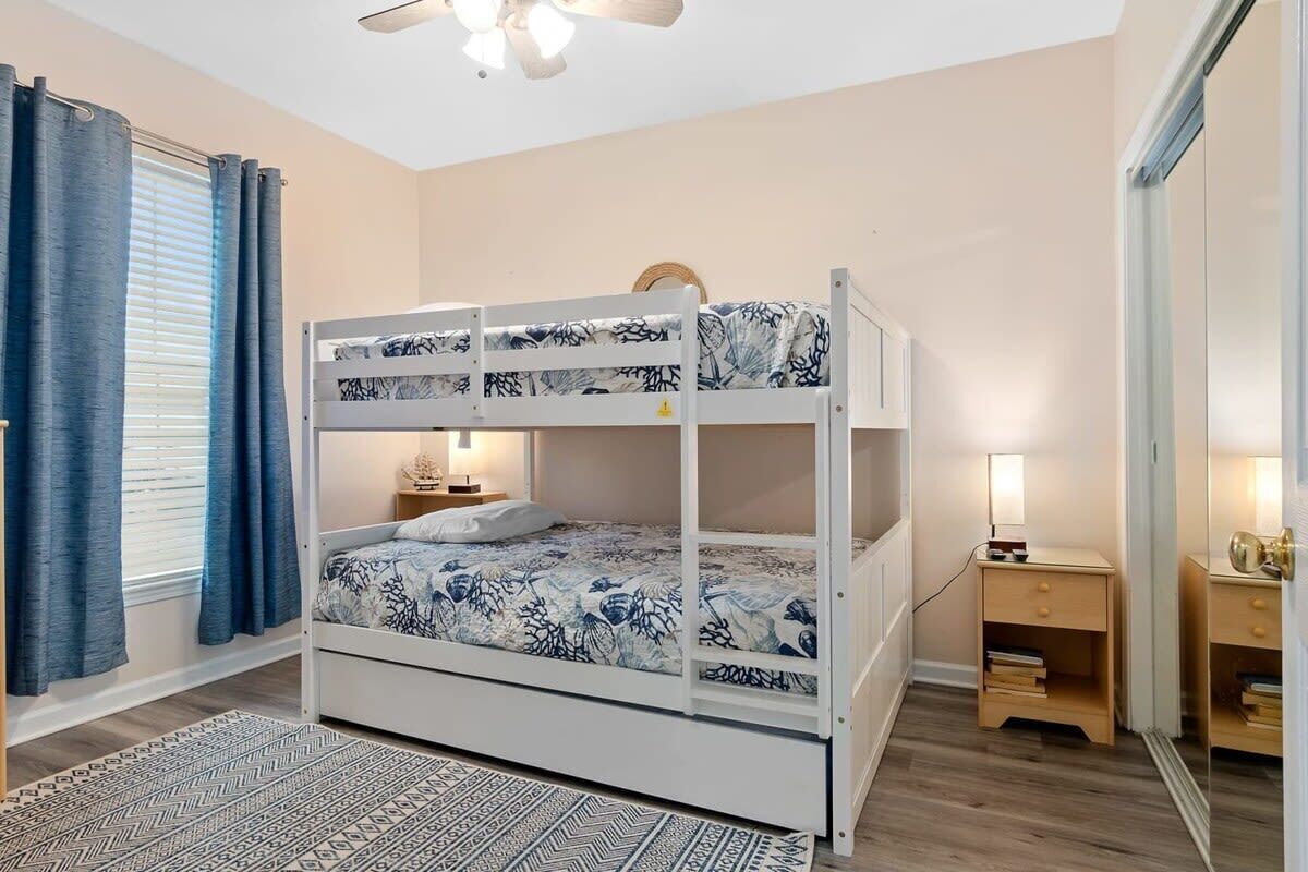 A bunk bed with a patterned bedspread, a bedside table with a lamp, a small wooden nightstand, and a patterned rug.